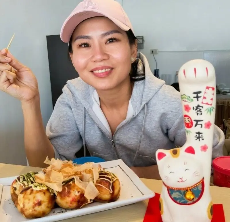 A smiling woman holding a skewer, with a plate of takoyaki and a lucky cat figurine on the table