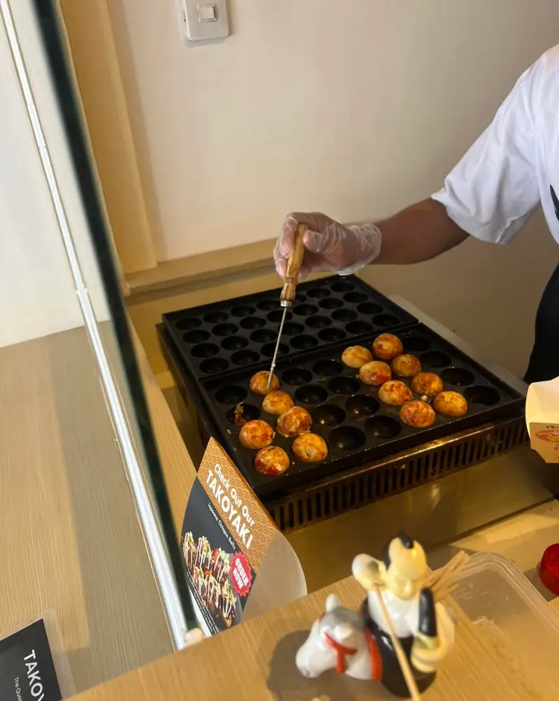 A cook in a plastic glove turning takoyaki with a pick on the hot griddle