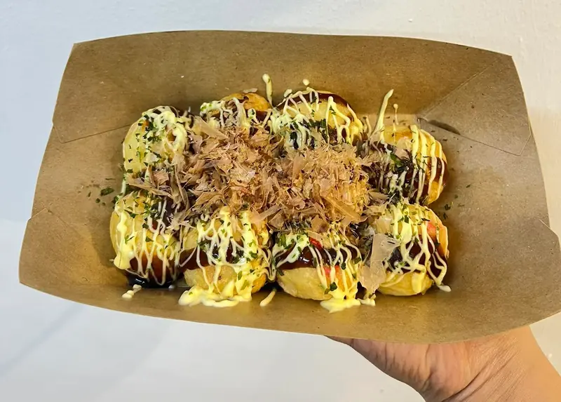A kraft box of takoyaki with mayo, sauce, nori and bonito flakes, held up by hand