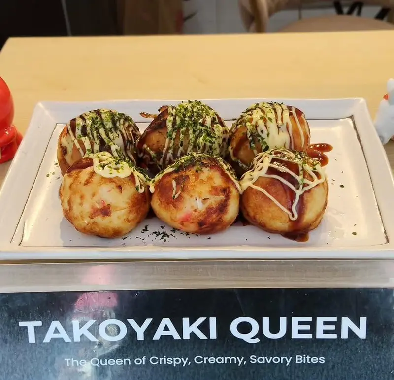 Six takoyaki with mayo, sauce and nori on a plate in front of the Takoyaki Queen sign